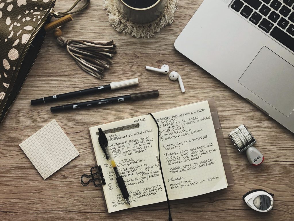 Top down view of notebook, laptop and stationery items on a wooden surface. Items include a green and white spotted pencil case with a tassel zipper pull, a stack of grid paper post-it notes, two tombow brush pens, airpods, small notebook opened to a page with random note-taking written in it, a date stamp, a small inkpad, the corner of a laptop and a coffee mug on a white macramé coaster.