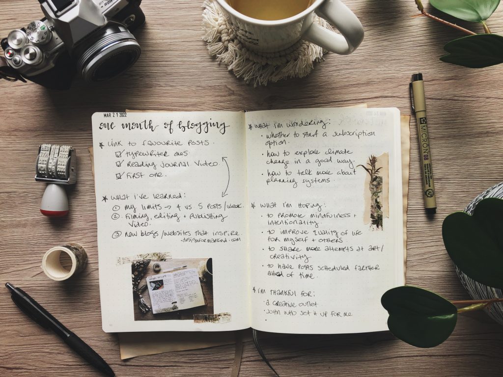 A top down view of a notebook with bogging notes, surrounded by stationery items. On the left of the notebook is a pen, a roll of washi tape, a date stamp and a vintage camera. At the top of the photo is a white mug of tea on a white macramé coaster. On the right is a micron pen and the edge of a houseplant.