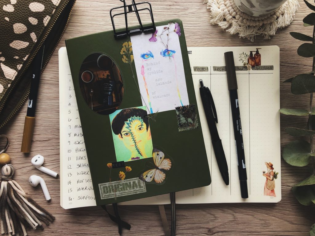 A top down view of notebooks on a wooden desk surface. The top notebook is closed with stickers decorating the cover. The notebook underneath it is open with some handwriting and stickers visible. To the left is a partial view of green and white spotted pencil case with a tassel zipper pull. A gold lidded black marker is seen in the opening. A set of airpods is right beside the notebooks. A black pen and black marker are set on top of the opened notebook page. At the top is the edge of a white macramé coaster. On the right is a stem of dried eucalyptus.