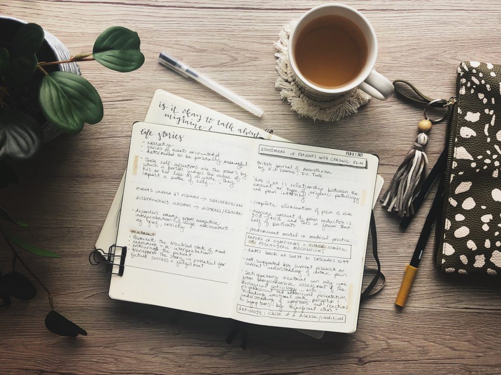 Notes from the Migraine World Summit. A top down view of the notebook on a wooden surface surrounded by a houseplant, a clear pen, a white mug of tea on a white macramé coaster, and a green and white spotted pencil case with a tassel zipper pull.