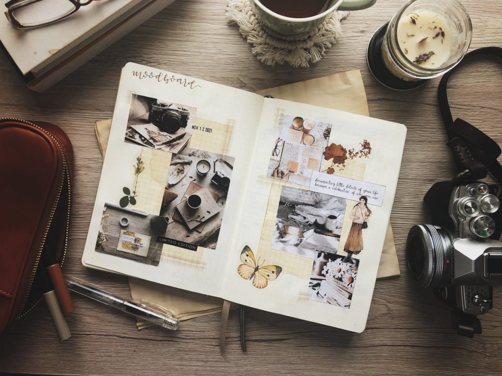 A topdown view of items on a wooden surface including a personal moodboard in a notebook. Elements of the moodboard are listed within the post. Other items in the frame include a tan leather pencil case, a stack of books and reading glasses, a mug of coffee on a white macramé coaster, a mason jar candle and a vintage camera.
