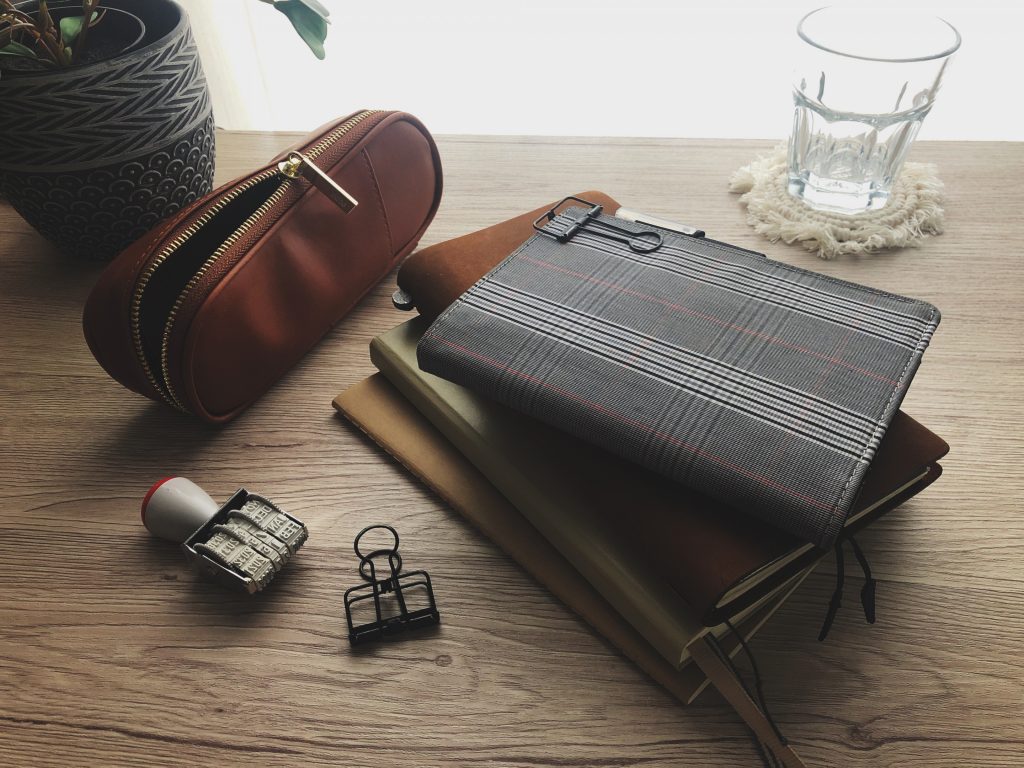 A stack of four assorted journals and notebooks on a wooden desk surface.  Behind the notebooks, a black and white patterned plant pot, a brown zippered pencil case and a glass of water on a white macramé coaster. In front of the notebooks is a black binder clip and date stamp.