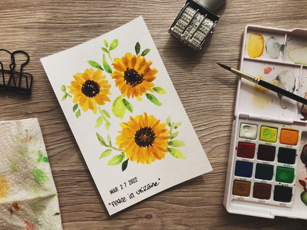 Painting sunflowers in solidarity for peace in Uknraine. On theright is a small watercolour palette with a paintbrush, Beside that a date stamp. In the centre a 4 X 6 card with three sunflowers painted on it. on the right a black binder clip and a piece of paper towel with paints blotted on it.