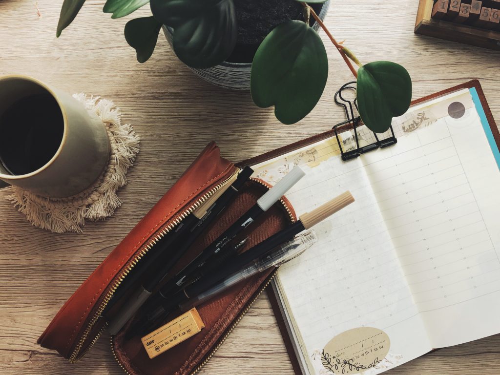 A top down view of journaling on a wood surface. on the left is a cup of coffee on a white macrame coaster. In the centre is a brown pencil case opened to show a wood rubber date stamp, a clear fountain pen and four neutral bush markers. On the right and partially under the pencil case is a traveler's notebook opened to a blank page decorated at the top and bottom left corner with neutral collage papers and tapes. The notebook is held open with a large skeleton clip at the top. Above the notebook is a partial view of some leaves from a houseplant.