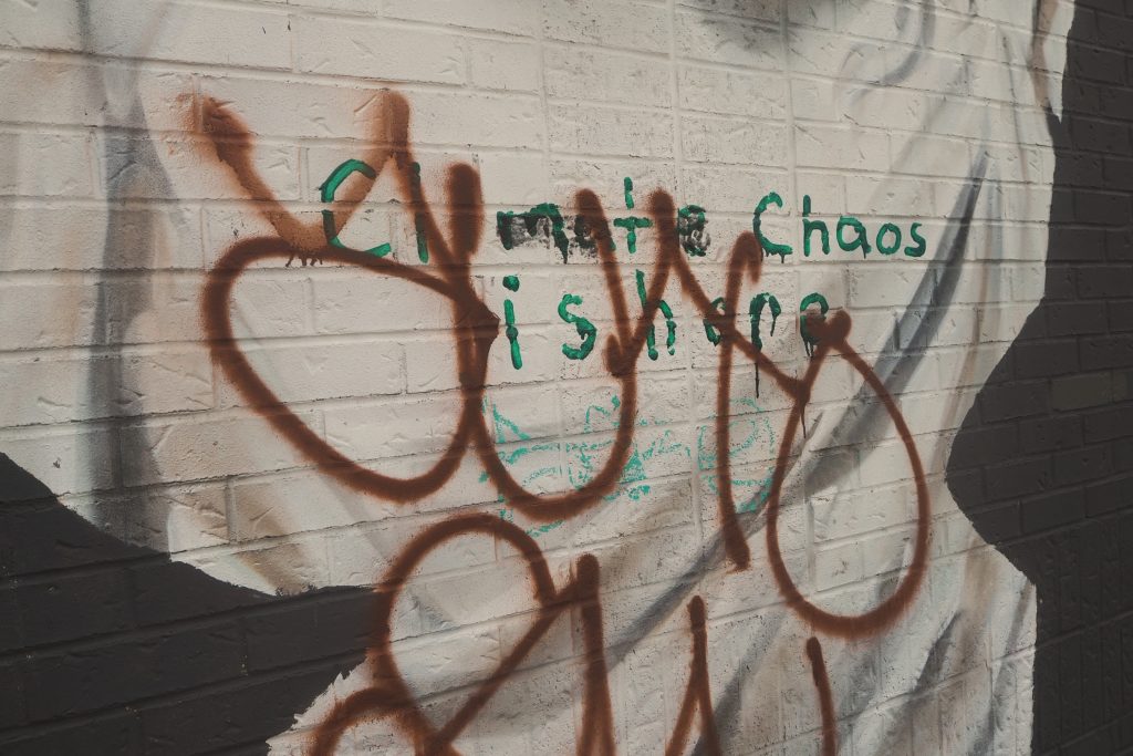 Graffiti on a white brick background says "Climate Chaos is here" in green with red lettering sprayed over it "YUXO"