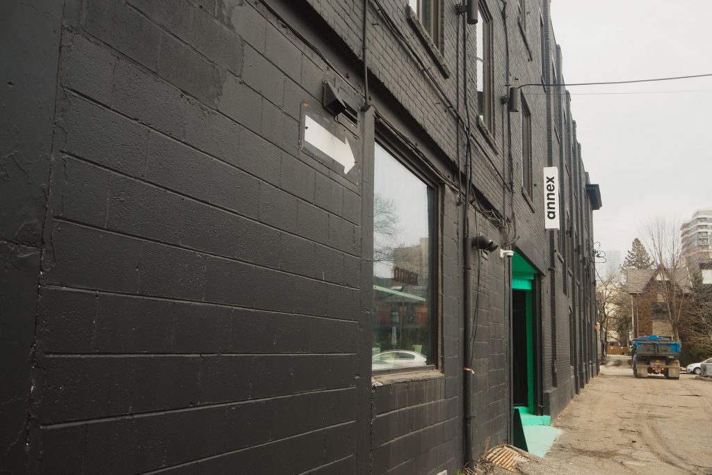 Angled view of a black painted building wall facing a narrow laneway. A white arrow painted on the building points forward. A white vertical sign above a doorway reads "annex". Pops of teal inside the doorway. In the distance a blue recycling truck is driving away.
