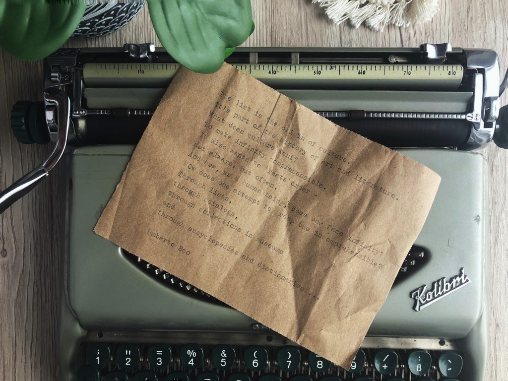 A top down view of a typewritten page set on a vintage typewriter. The page is a creased sheet of kraft packaging paper. The typewritten text is titled The List Is The Origin Of Culture and is contained in the body of blog post. The background of the photo is a wooden desktop and part of a houseplant can be seen in the top left corner.