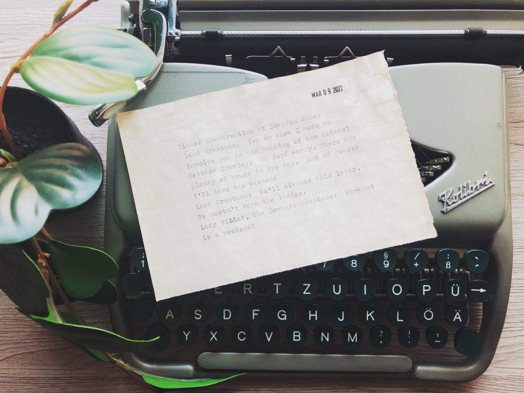 A half-sheet of coffee-dyed paper with typewritten text (the text is written out in full in the body of the post) sits on top of a vintage green Kolibri typewriter. A green houseplan is partially visible on the left. The partially visible table top is light wood grain.