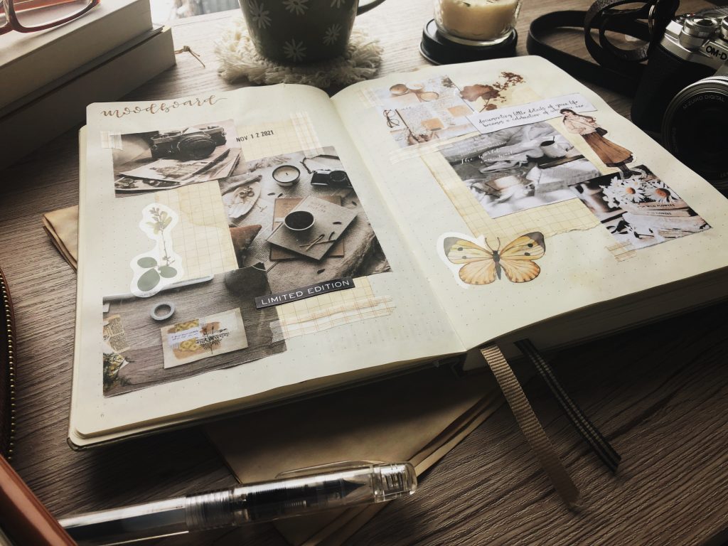 A notebook laying open on a wood surface. The pages are decorated with aesthetic photos of coffee, candles, books and flowers, and stickers. The notebook is surrounded by a clear fountain pen, a vintage camera, a small jar candle, and a stack of two books. 