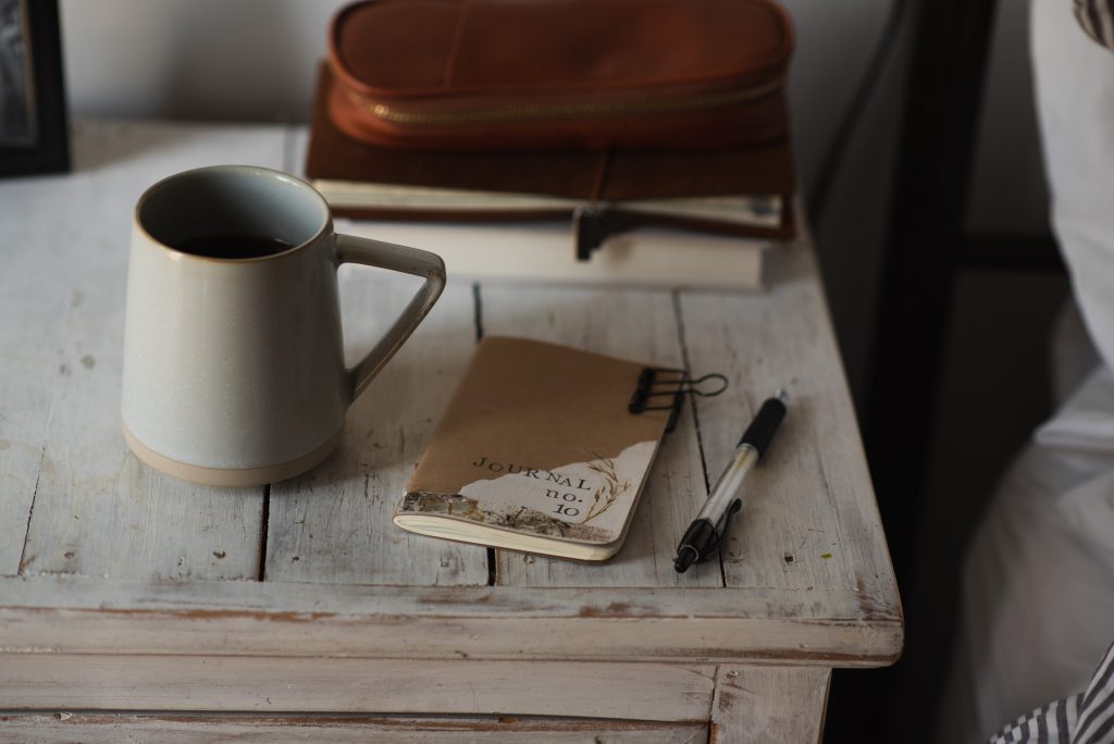 A Morning Pages journal,, ballpoint pen and coffee mug on a white rustic bedside table.