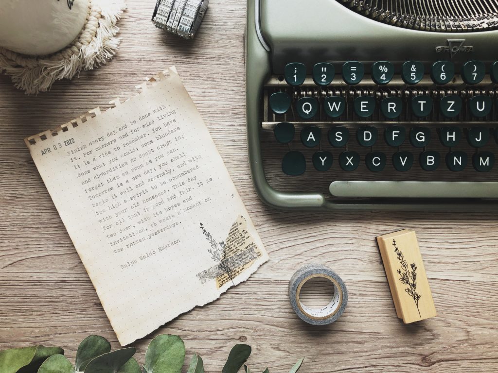 On a light wood backdrop, a sheet of paper torn from a notebook with a typewritten paragraph: "Finish every day..." The text is in the body of the post. Above the paper are the edge of a white mug on a white macramé coaster and date stamp. At the top right is a vintage green typewriter. Below it are a roll of grey washi tape and a wooden stamp with a floral stem image. A partial piece of eucalyptus leaves at the bottom left.