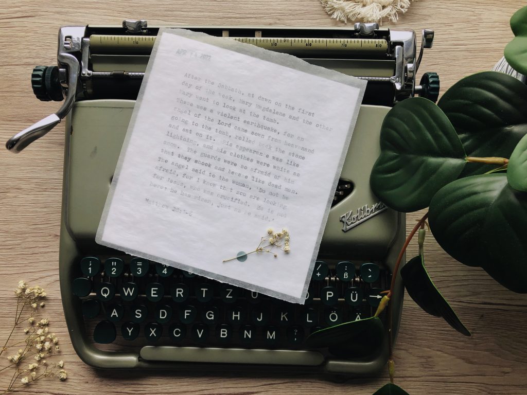 A birds-eye view of a vintage typewriter with a square piece of typed vellum laid on top. Vellum is decorated in the bottom corner with a piece of dried baby's breath. The backdrop is light  wood. Part view of a green houseplant on the right side. Typewritten text is the Easter story, printed in full in the body of the post.