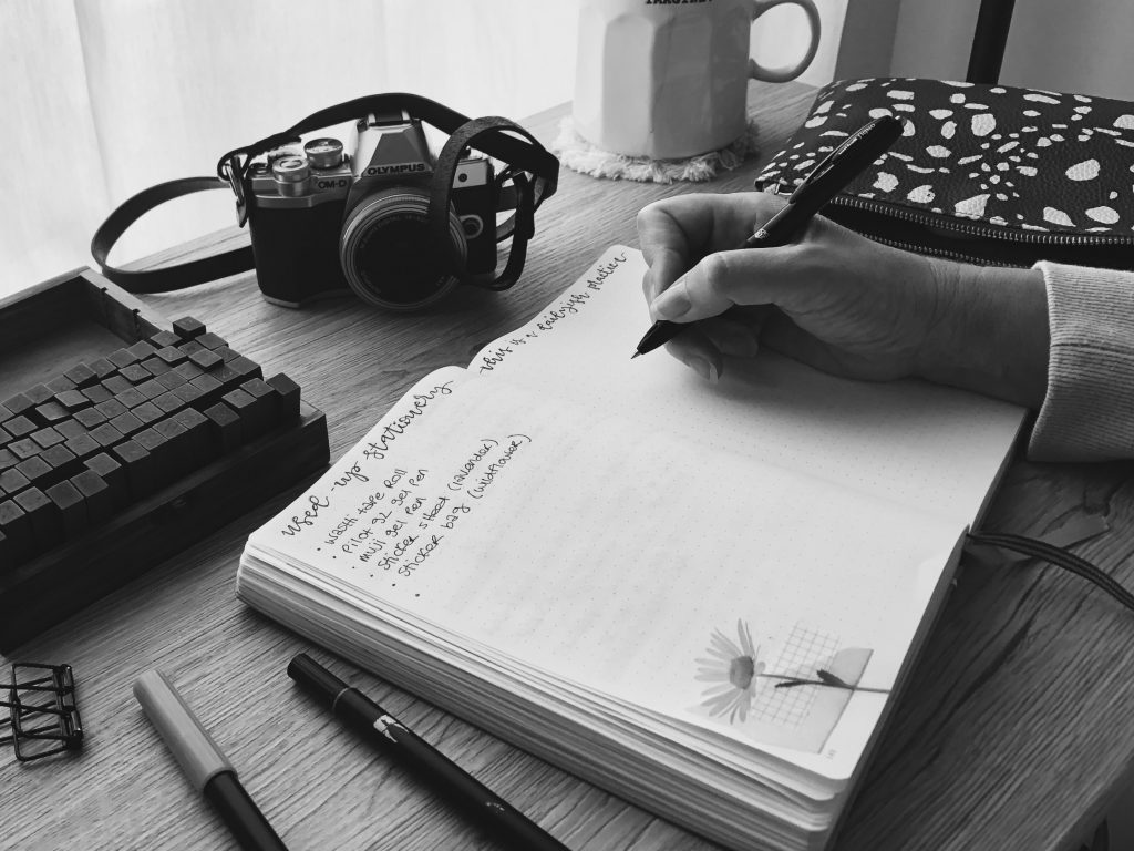 A black and white photos depicting a daily writing practice. A hand holding a pen over the blank page of a notebook. Also on the desk, two markers, a box of wooden alphabet stamps, a vintage camera, a coffee mug and a pencil case.