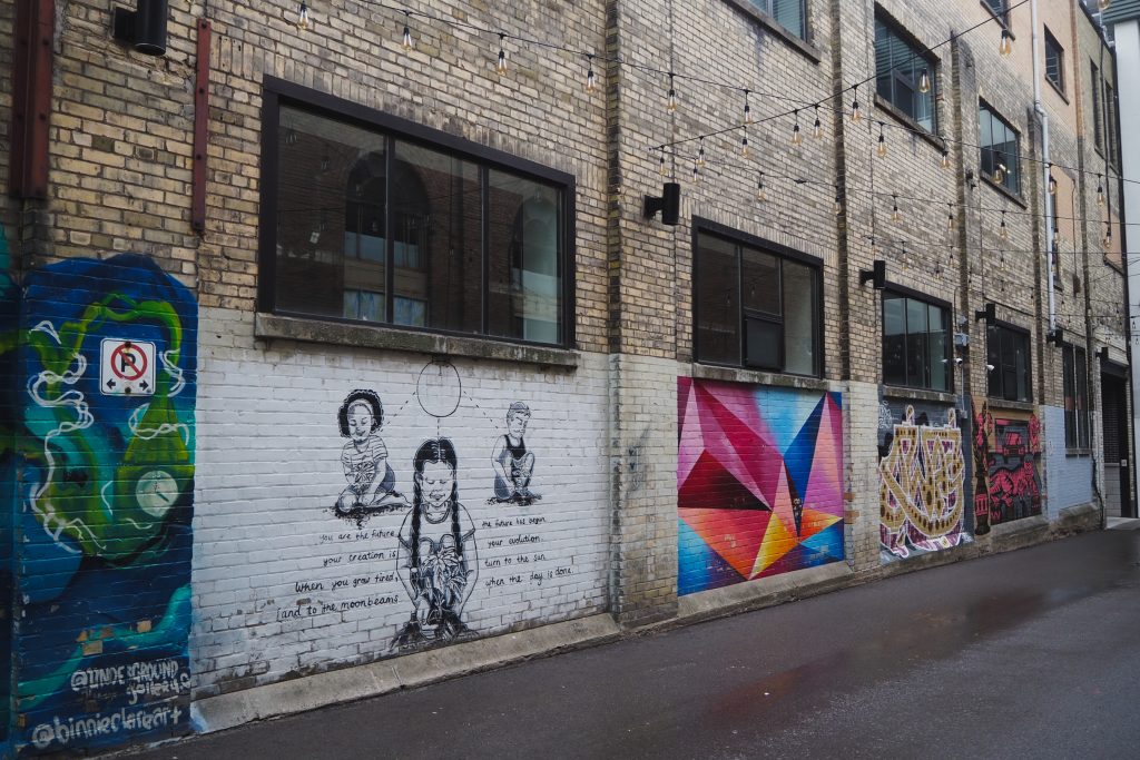 Street photography in Kitchener Ontario. A row of street art is painted on the lower part of a brick wall in an alleyway.