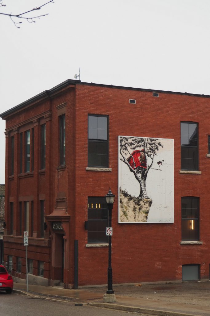 Street photography in Kitchener, Ontario. A red brick low rise buidling with a mural on its side depicting a whimsical cliff with a tree and treehouse on its edge, and a diving board out the side with a diver poised on the end of it.