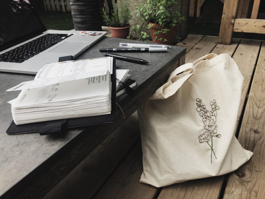 A photograph of a portable work space. A low coffee table on a an outdoor deck with an open notebook, a laptop and a few pens scattered on it. A floral embroidered tote bag leans against the the table.