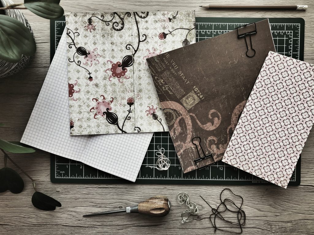 A photograph depicting the supplies and stages of making my own notebooks. There are four notebooks in various stages of completion, with patterned paper covers, set on a cutting mat. around the mat are an awl, several colours of embroidery floss and a pencil.
