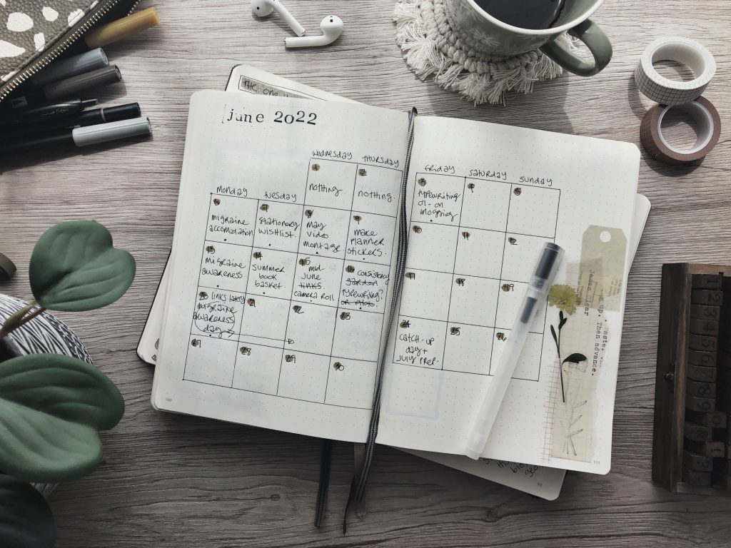 A view of an open notebook showing a June calendar layout. Also on the desk, the corner of a pencil case with markers spilling out, a coffee mug on a white macramé coaster, two rolls of washi tape, a houseplant, and a wooden alpha-numeric stamp set.