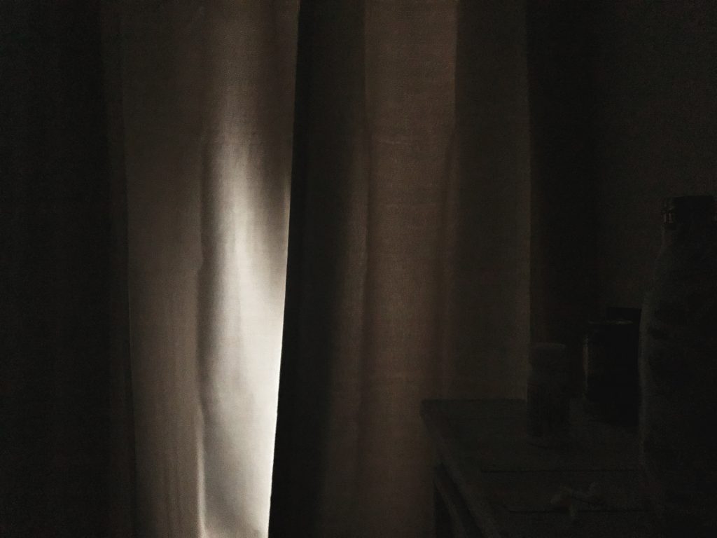 A set of closed room-darkening drapes with a shaft of light between the panels. It represents the light of an act of kindness in a dark time.