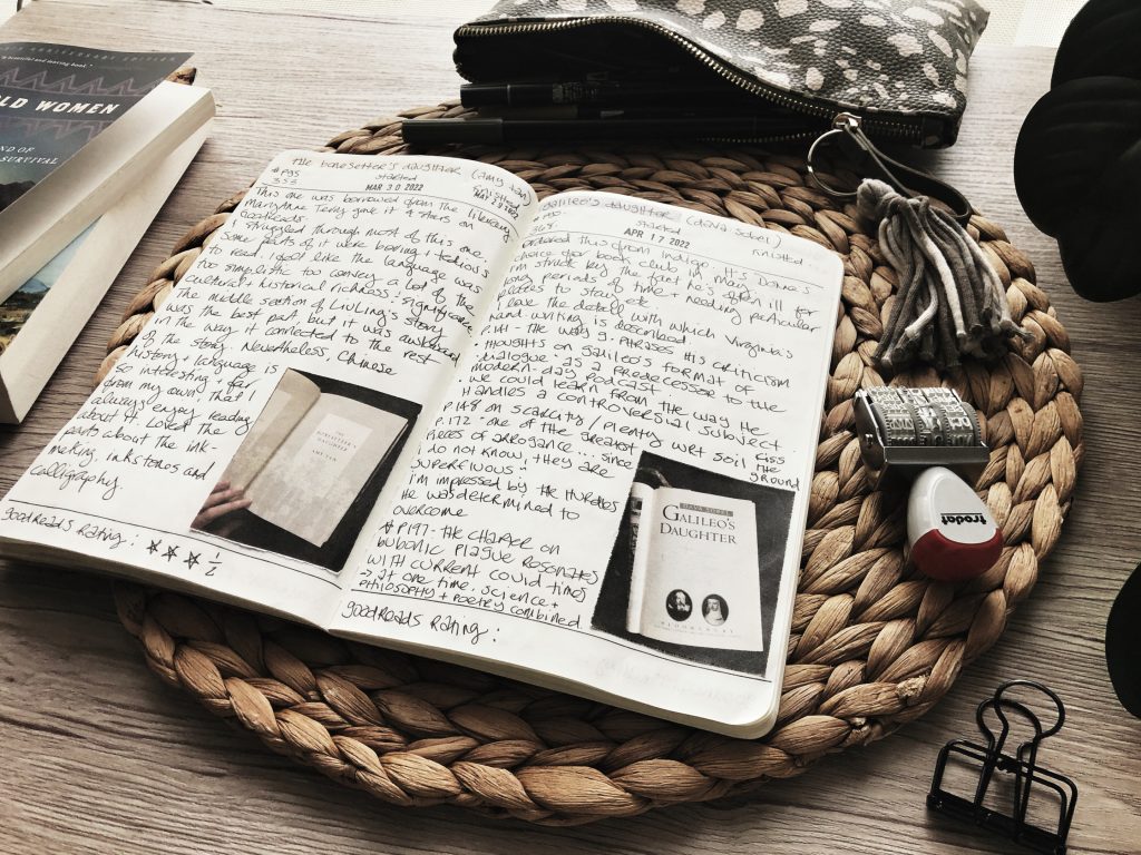 An open notebook with writing across the pages, and two photos of book covers pasted in. The notebook is set on a round raffia placemat on a wooden desk surface, along with various small stationery items. the edges of a stack of two books is visible to one side.