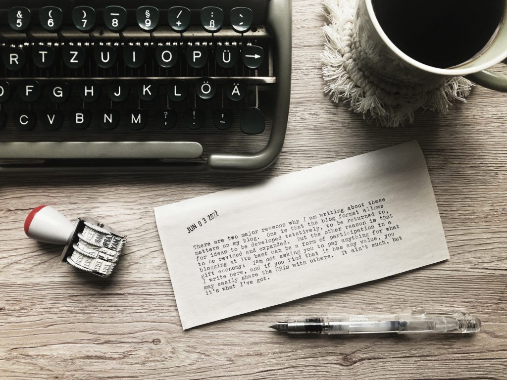 A top-down view of a typewritten half-sheet of newsprint tucked under the corner of a vintage typewriter. Also on the desk: a date stamp, a fountain pen, and a green mug of coffee on a white macramé coaster. The text on the paper is contained within the post.