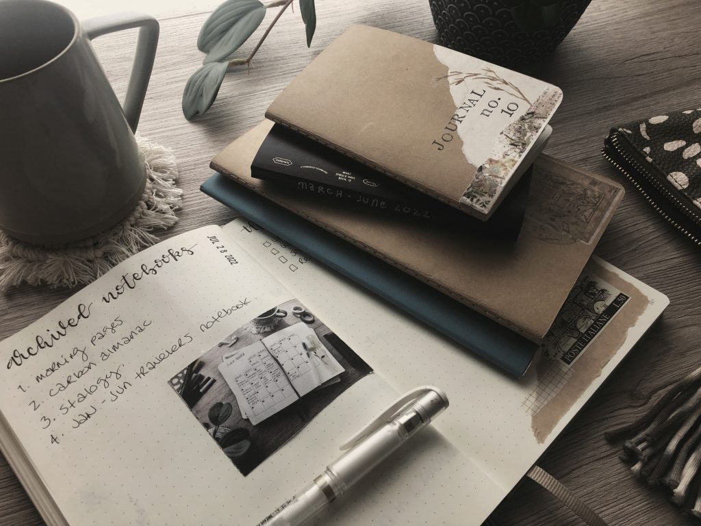 A photo depicting my recently archived notebooks. There are four notebooks stacked on one side of my opened blogging notebook on my desk. Also on the desk re a coffee mug, houseplant and pencil case.