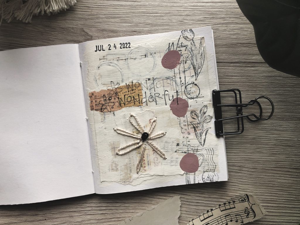 A photograph of how I'm using one of my mini sketchbooks. A small square handmade notebook is opened at a page with collage elements pasted, painted and stitched onto it.
