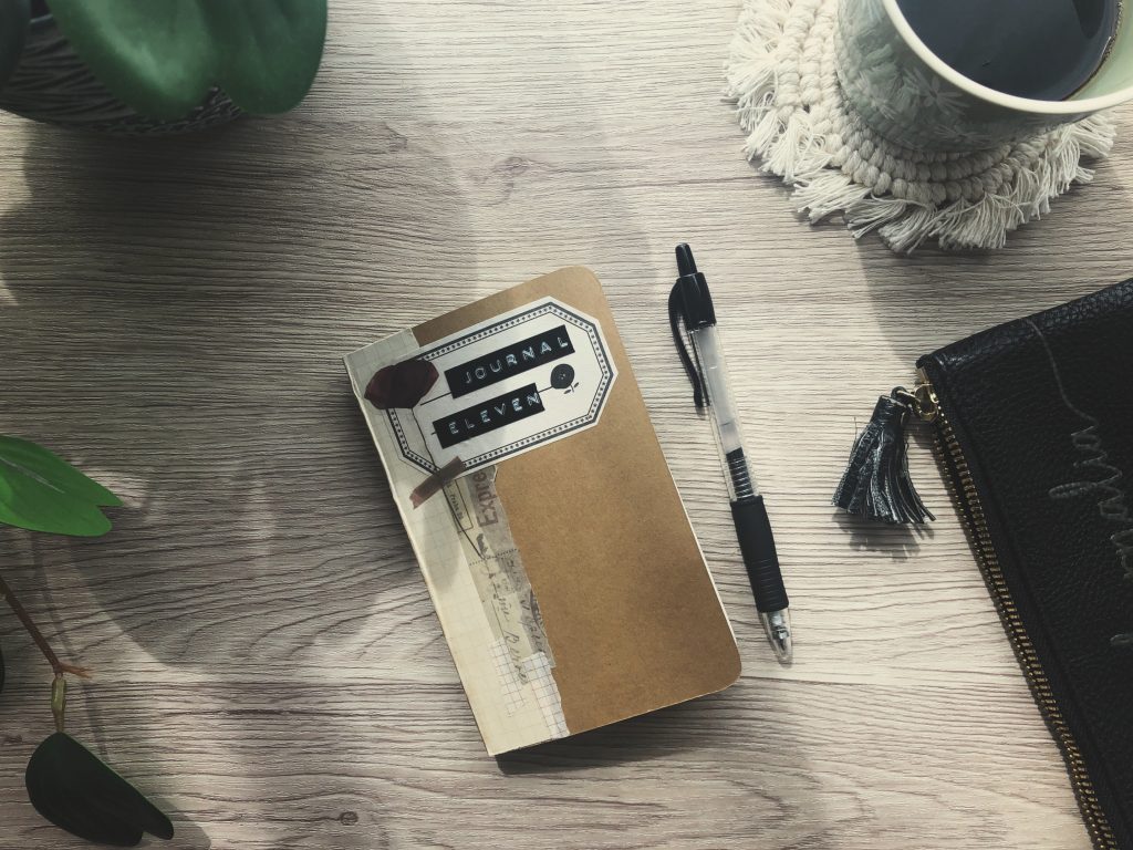 A photo depicting morning pages journaling. A small moleskine, the cover decorated with collage papers and a floral sticker sits on a wooden desk surrounded by a pen, pencil case, coffee mug and green plant.