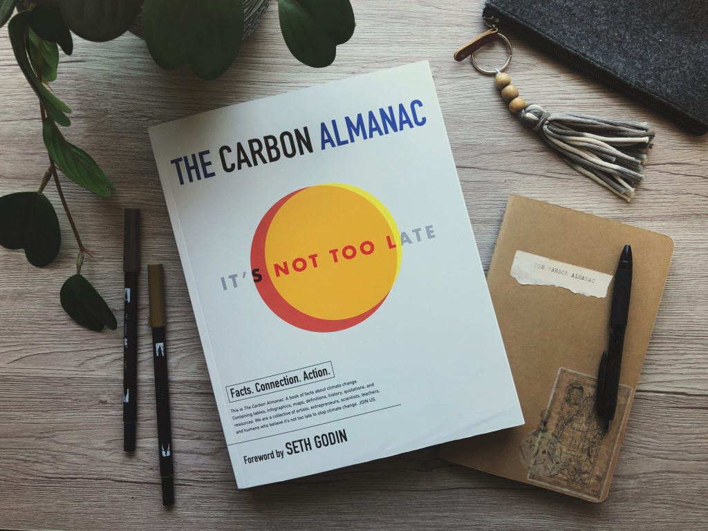 A photograph of The Carbon Almanac on a wooden desk surface, surrounded by pens, a notebook, pencil case and a houseplant.