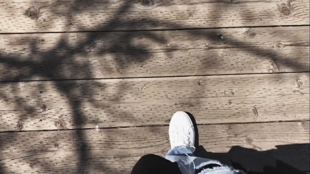 An outtake from the Carbon Almanad Launch video. A photo looking down at oneself walking in white sneakers and distressed jeans on a boardwalk surface.