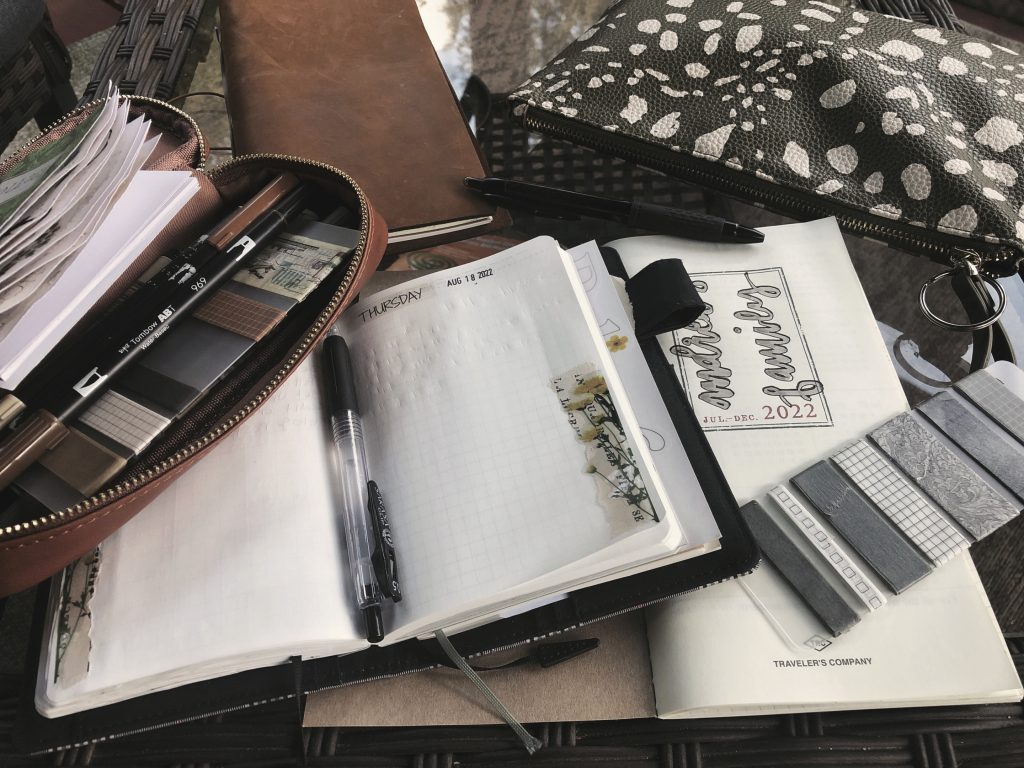 A photo depicting journaling away from home. Several notebooks, pencil cases and stationery supplies are spread out across an outdoor table.