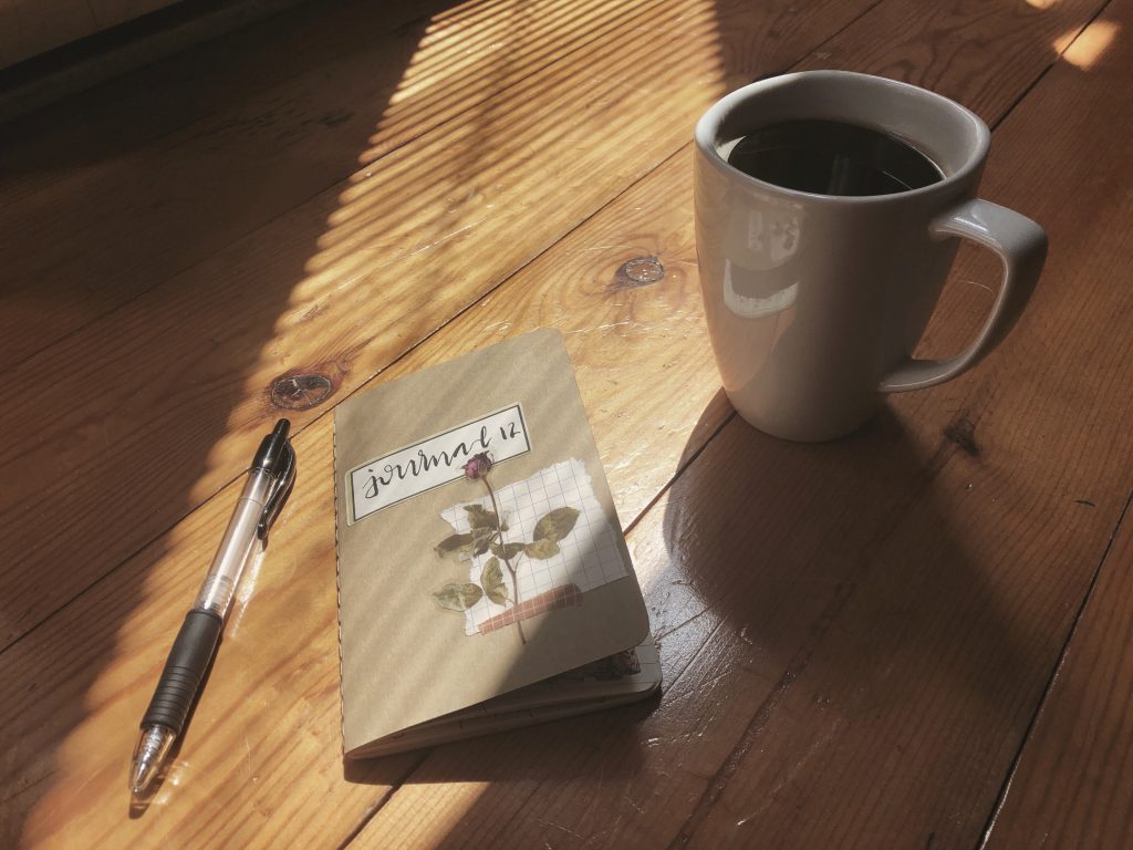 A photograph representing this morning's gratitude journaling. There is a wide shaft of light on a golden pine wood surface. In the light there is a pen, a notebook decorated with a floral sticker and a hand-lettered label that reads Journal 12, and a white mug filled with coffee.