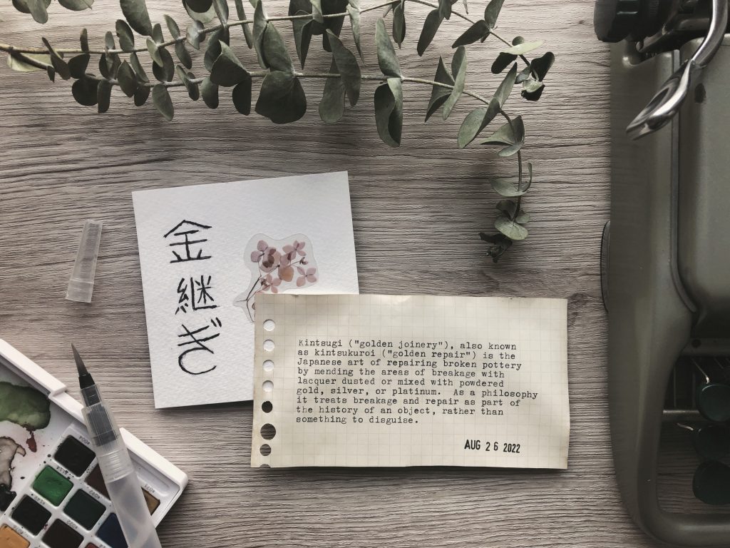 A photo displaying typewritten text (written in the body of the post) along with a square card depicting the Japanese kanji 金継ぎ painted with black watercolour. Also on the desk, a paint palette and brush, a vintage typewriter and sprigs of dried eucalyptus.