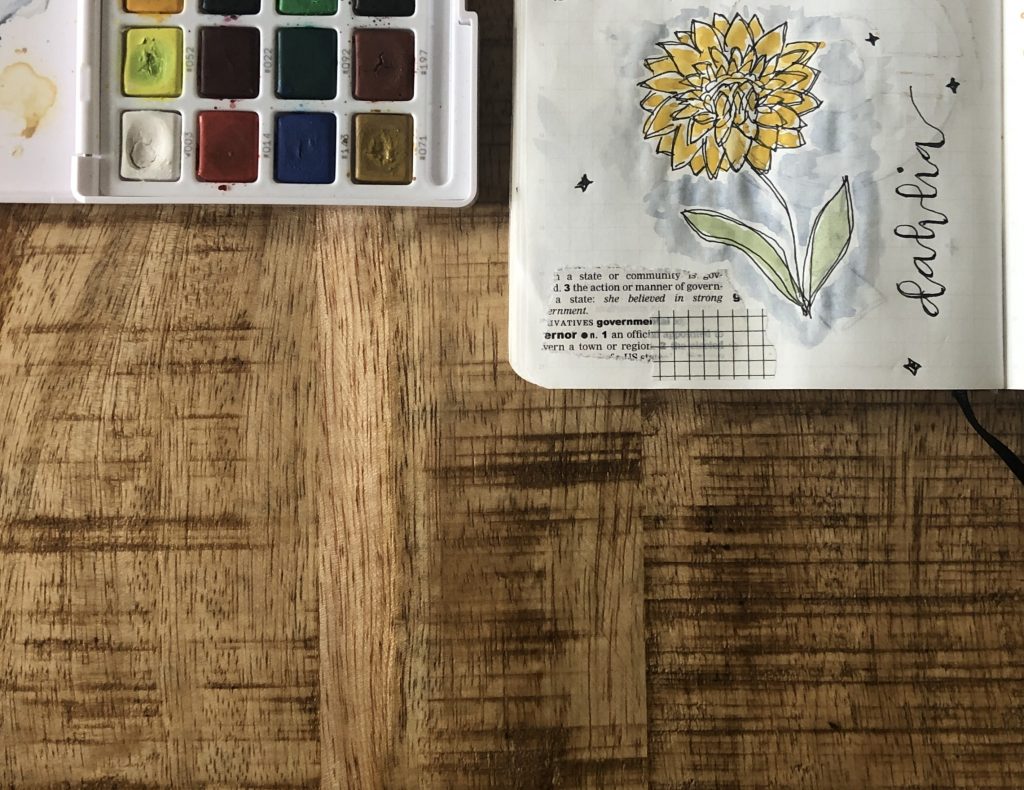 A little watercolour painting of a dahlia on a page in my daily planner. Beside the planner is an open watercolour palette.