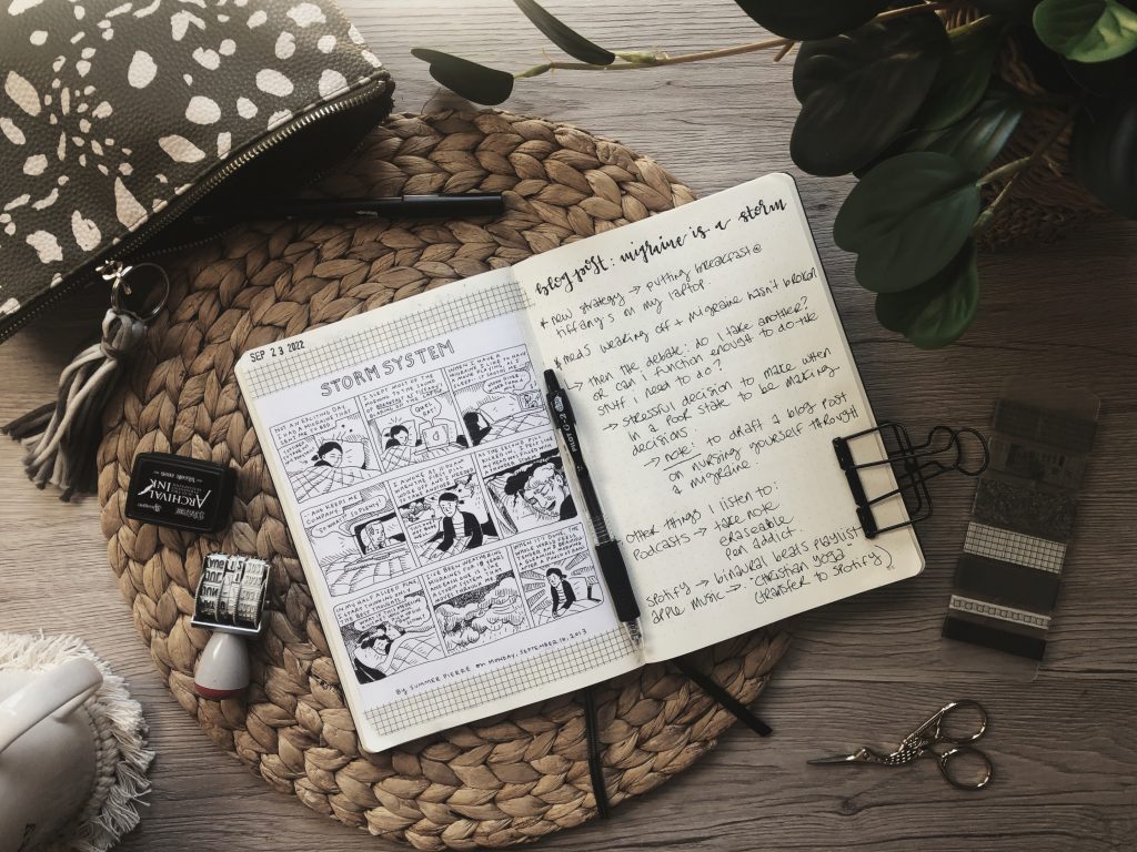 A photo of an open notebook with the comic strip "Storm System" pasted on the left side, and blogging notes written on the right. Also on the desk, various stationery supplies, a coffee mug and a houseplant.