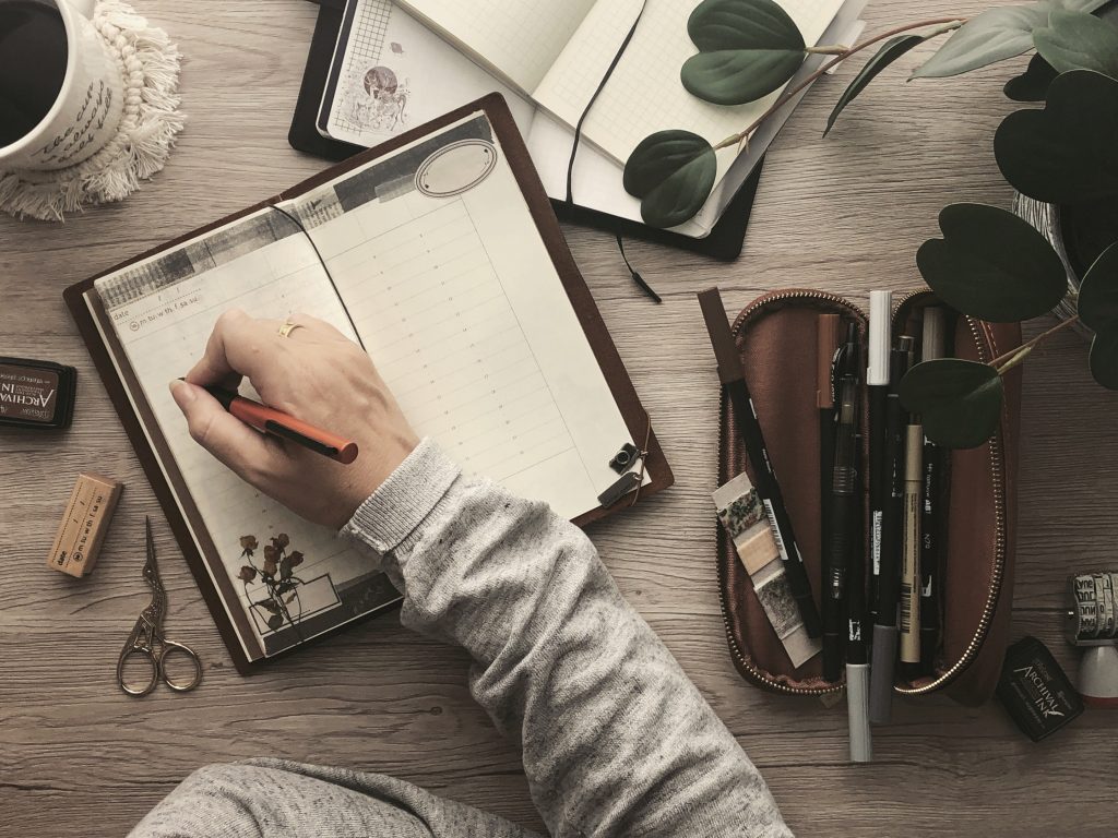A photo depicting taking time to reset. A hand, holding a pen, hovers over a blank journal page. Various stationery items such as a pencil case and scissors, also a coffee mug and houseplant, are scattered around the desk.