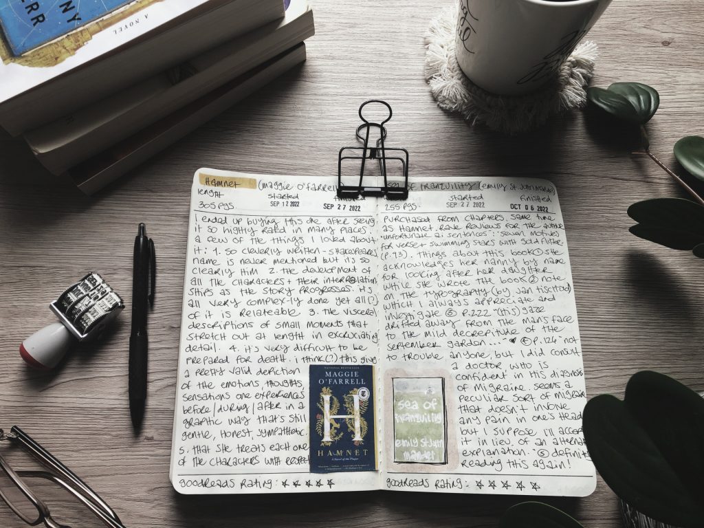 A quick fall reading update. The pages of a reading journal are opened to the handwritten reviews of Hamnet and Sea of Tranquility. Also on the desk, a green houseplant, reading glasses, a coffee mug, a stack of paperbacks, a pen and a date stamp.