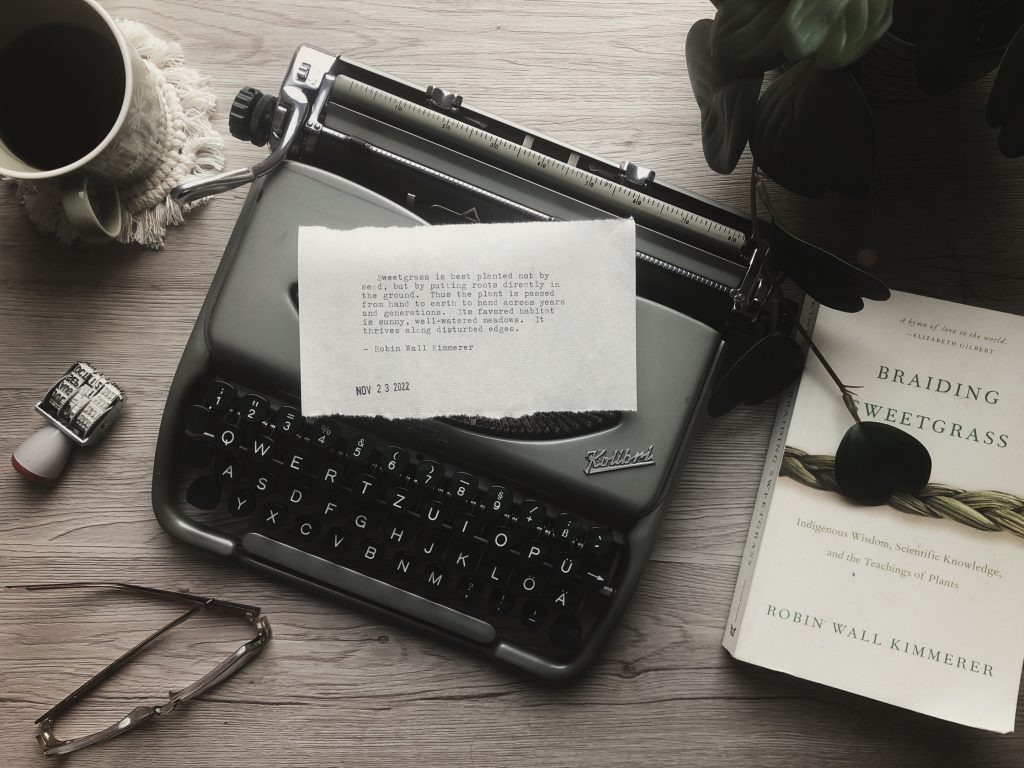 Typewritings 14: Facts about sweetgrass. A wooden desktop with a vintage typewriter, a coffee mug, houseplant, and a copy of the book "Braiding Sweetgrass". On the typewriter is a typewritten quote from the book, the text of which is copied into the post.